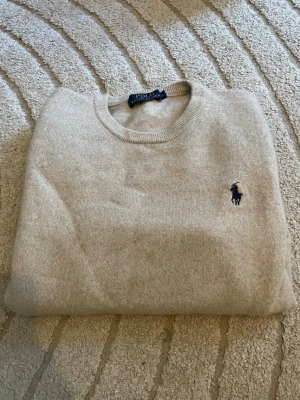 100% Merinoull Beige stickad tröja från Polo Ralph Lauren - Säljer en beige stickad tröja från Polo Ralph Lauren 100% Merinoull med rund halsringning och den klassiska mörkblå logotypen broderad på bröstet. Tröjan har långa ärmar och ribbade muddar vid ärmslut och nederkant. Perfekt för dig som gillar stilren och tidlös design. Strl L men krymt i tvätten så passar xs/s. Därav priset