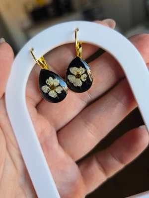 Svart droppformade örhängen med blommor - Unika örhängen i droppform med svart bakgrund och inneslutna vita blommor. Örhängena har guldfärgade krokar och en elegant, handgjord känsla. Perfekta för dig som gillar naturinspirerade accessoarer och vill sticka ut med något personligt.