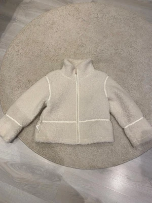 Vit teddyjacka med hög krage - Mysig beige teddyjacka med hög krage och dragkedja framtill. Jackan har markerade sömmar i vitt och raka ärmar. Perfekt för att hålla värmen och ge en trendig look. Materialet är fluffigt och mjukt, vilket gör jackan superbekväm.