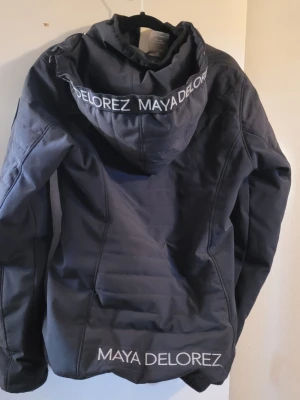 Svart vindjacka från Maya Delorez - Snygg svart vindjacka från Maya Delorez med huva och dragkedja framtill. Jackan har två fickor med dragkedja och logga på bröstet, ryggen och huvan. Materialet är lätt och vindtätt, perfekt för blåsiga dagar.