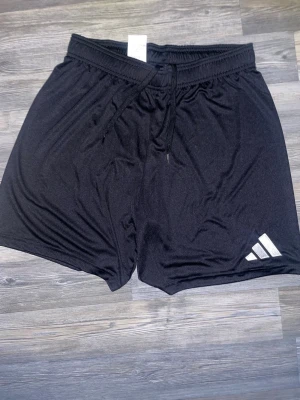 Svarta träningsshorts från Adidas - Säljer ett par svarta träningsshorts från Adidas med klassisk logga på vänster ben. Shortsen har elastisk midja med snörning och är tillverkade i ett lätt och snabbtorkande syntetmaterial. Perfekta för gymmet eller löprundan.