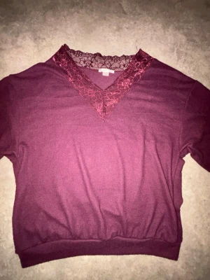 Vinröd stickad tröja med spetsdetalj - Mysig vinröd stickad tröja med v-ringning och dekorativ spets i samma färg runt halsen. Tröjan har långa ärmar och ribbade muddar vid nederkant. Perfekt för dig som gillar en feminin touch på klassiska stickade plagg. 