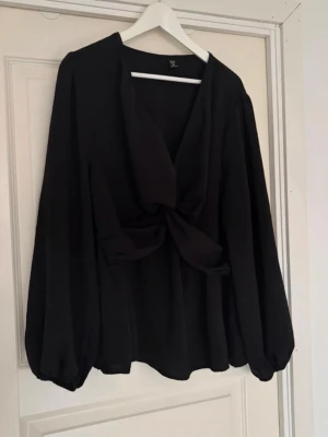 Svart blus med knyt framtill - Stilren svart blus med v-ringning och knyt framtill. Blusen har långa, puffiga ärmar med resår vid ärmsluten och en lös passform. Perfekt för dig som gillar en enkel men trendig look. 3XL, nyskick använd någon gång 