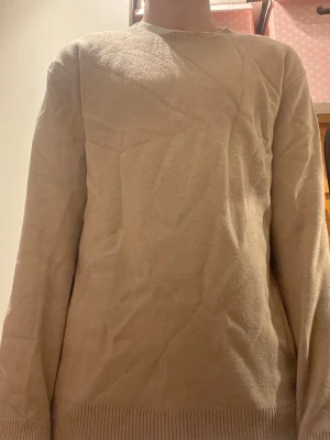 Beige stickad tröja med rund hals - En enkel och stilren beige stickad tröja med rund hals och långa ärmar. Tröjan har ribbstickade muddar vid ärmslut och nederkant. Perfekt basplagg för lager på lager och passar till det mesta. 