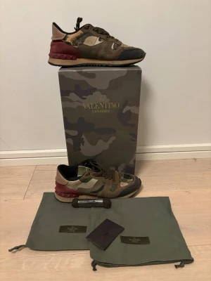 Valentino rockrunners  - Säljer ett par Valentino Garavani Rockrunner sneakers i camouflage med inslag av grönt, brunt, beige och vinrött. Skorna har snörning, platt sula och detaljer i mocka och skinn. Kommer med originalkartong, dustbags och extra skosnören. Stl 38 i ett bra skick
