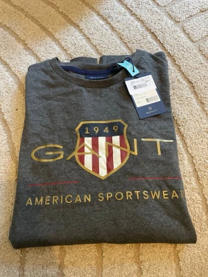 Grå långärmad t-shirt från GANT - Snygg grå långärmad t-shirt från GANT med stort tryck framtill i form av ett sköldmärke och texten 'American Sportswear'. T-shirten har rund hals och är tillverkad i mjuk bomull, perfekt för en avslappnad och sportig stil.