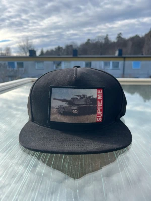 Svart Supreme keps, tank, SS20 - Säljer en svart snapback-keps från Supreme med ett stort patch framtill som visar en stridsvagn och röd SUPREME-text. Autentisk och i nyskick.
