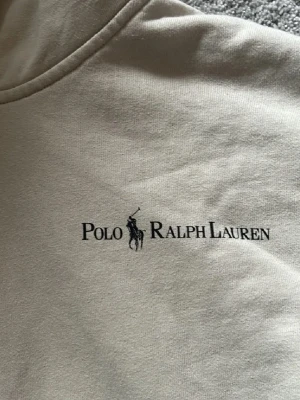 Beige hoodie från Polo Ralph Lauren - Snygg beige hoodie från Polo Ralph Lauren med svart logga och klassisk känguruficka. Hoodien har dragsko i huvan och är tillverkad i mjukt bomullsmaterial. Perfekt för en avslappnad och stilren look.