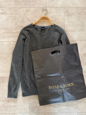 Rose & Born tröja  - En stilren mörkgrå/mörkgrön tröja från Rose & Born med rund halsringning och långa ärmar. Tröjan har ribbade muddar vid ärmslut och nederkant. Perfekt för dig som gillar en clean och klassisk look.