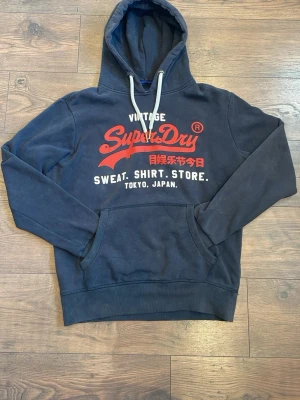 Mörkblå Superdry hoodie med tryck - Mörkblå hoodie från Superdry med stor röd och vit logga på bröstet samt japansk text. vita dragsnören i huvan och mjukt material. Perfekt för dig som gillar streetwear och vill ha en snygg och bekväm hoodie. Hoodin är använd mycket så det finns kännetecken på användning, hoodin har en fläck på armen som finns på bilderna.