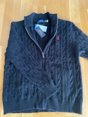 Ralph Lauren - Snygg svart kabelstickad tröja från Polo Ralph Lauren med halv dragkedja och ribbad krage. Tröjan har det klassiska röda Polo-logot broderat på bröstet och ribbade muddar vid ärmslut och nederkant. Perfekt för lager på lager och stilren look.