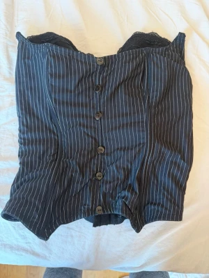 Svart randig korsett med knappar - Snygg svart korsett med tunna vita ränder och knäppning framtill. Modellen är axelbandslös och har spetsdetaljer upptill. Materialet är troligtvis bomullsmix med inslag av stretch för en bekväm passform. Perfekt för att skapa en cool och stilren look. i storlek s!