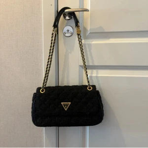 Väska från Guess - Snygg svart quiltad handväska från Guess med guldfärgade kedjeremmar och klassisk trekantslogga framtill. Väskan har en elegant form och axelrem i kombinerat skinn och kedja, vilket ger en lyxig känsla. Perfekt accessoar för att lyfta din outfit.