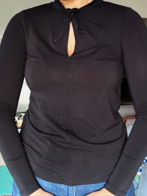 Svart långärmad topp med knyt - Snygg svart långärmad topp med knyt i halsen och en liten öppning framtill. Toppen har en slim passform och är tillverkad i ett mjukt, stretchigt material som känns bekvämt mot huden. Perfekt att styla med jeans eller kjol för en clean look. Står storlek XS men jag skulle säga passar mer S/38/M