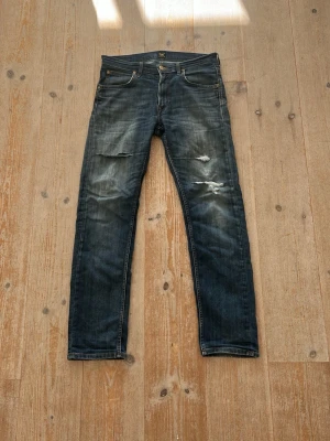 Blå slitna jeans från Lee - Säljer ett par blå jeans från Lee med slitningar och hål på låren och knäet. Jeansen har klassisk femficksmodell, raka ben och tydliga tvättade detaljer för en cool, avslappnad look. Perfekta för dig som gillar en trendig och casual stil.