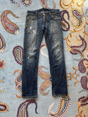 Blå slitna jeans från Dsquared 2 - Säljer ett par blå jeans från Dsquared 2 med slitningar och distressed detaljer på både fram- och baksida. Trådarna i slitningarna har gått av på vissa ställen som syns på bilderna, men i övrigt i fullt godkänt skick. Inköpta begagnat i Paris 2022 för drygt 2500kr.