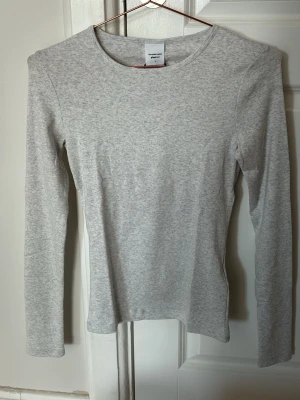 Ljusgrå långärmad topp Gina Tricot - En enkel och stilren ljusgrå långärmad topp från Gina Tricot, Elevated Basics-kollektionen. Toppen har rund halsringning och är gjord i mjuk bomullsmix som känns skön mot huden. Perfekt att matcha med jeans eller kjol för en clean look.