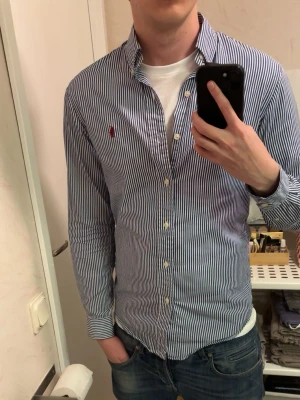 Ralph Lauren Skjorta - Säljer denna snygga slim-fit skjorta från ralph lauren! Perfekt nu till våren och sommaren💯Skjortan har ett trendigt randigt mönster och kommer i en snygg blå färg. Passar både att ha på med en tshirt under som med en tröja ovanpå! Modellen är 186 ish så den passar nog bättre på någon lite kortare än det. Tveka inte på att skriva om du har några frågor eller funderingar🙏