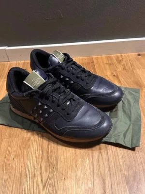 Svarta Valentino Garavani sneakers - Snygga svarta sneakers från Valentino Garavani med ikoniska nitar på sidorna. Skorna har en mix av läder och mocka, svarta skosnören och en beige sula. Tungan har en grön logotypdetalj. Perfekta för dig som vill ha en edgy och exklusiv look.