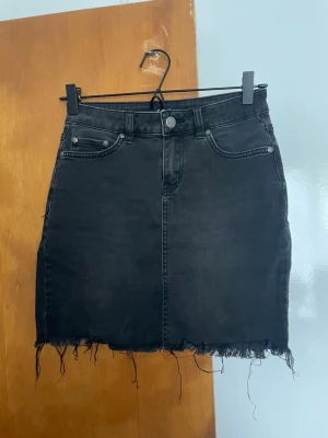 Svart jeanskjol med rå kant - Säljer en svart jeanskjol i klassisk femficksmodell med hög midja och rå, fransig kant nedtill. Kjolen har knapp- och dragkedjestängning samt bälteshällor. Perfekt för en avslappnad och trendig look.