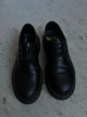 Dr. Martens 1461 Bex - Säljer ett par klassiska svarta loafers från Dr. Martens i äkta läder. Skorna har rund tå, grov sula med gul söm och snörning framtill. Perfekta för dig som gillar en chunky och tidlös stil.