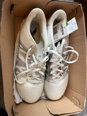 Vita och grå fotbollsskor från Adidas 44 nya - Snygga fotbollsskor från Adidas i vitt och grått med klassisk design och snörning. Skorna har en sula med små dobbar som ger bra grepp på konstgräs. Perfekta för dig som vill ha stilrena och funktionella skor på planen.