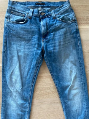 Blå jeans från Nudie Jeans - Säljer ett par klassiska blå jeans från Nudie Jeans med fem fickor och snygga slitningar framtill. Jeansen har en smal passform och ikonisk brodyr på bakfickorna. Perfekta till en avslappnad stil. 
