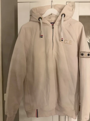 Vit hoodie från  Who’s A.C.E med dragkedja - Snygg beige\vit  hoodie från who’S  A.C.E. Den är använd 2 gånger och är bra material . Har  hel dragkedja, broderad logga på bröstet och cool detalj på ärmen. Huva med dragsko och ribbade muddar. Fodrad insida och detaljer i rött, vitt och blått på snören och krage. Perfekt för en avslappnad streetstyle.