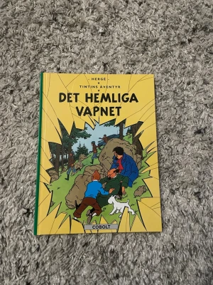 Tintins äventyr: Det hemliga vapnet - Säljer seriealbumet 'Det hemliga vapnet' från Tintins äventyr av Hergé. Boken har ett färgglatt omslag med illustrationer av Tintin, Kapten Haddock och Milou. Hårdpärm i gult, grönt och blått. Perfekt för dig som gillar klassiska serier och äventyr.