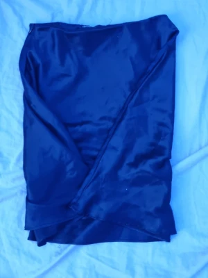 Mörkblå omlottkjol i satin - Snygg mörkblå omlottkjol i glansigt satinliknande material. Kjolen har en stilren design med dragkedja bak och omlott-detalj framtill. Perfekt för dig som gillar en elegant och trendig look.