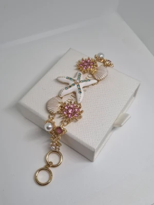 Guldigt armband med sjöstjärna och pärlor - Unikt armband i guldton med vita snäckor, en vit sjöstjärna dekorerad med turkosa stenar och rosa blommor med glittrande stenar. Armbandet har även vita pärlor och detaljerade länkar. Perfekt för dig som gillar havsinspirerade accessoarer.