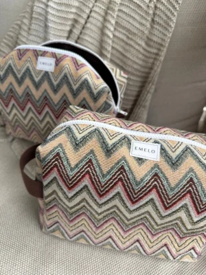 Necessärer med zigzag-mönster från Emelo - En necessärer i tyg från Emelo med färgglatt zigzag-mönster i beige, rött, grönt och blått. Den har vit dragkedja och en praktisk brun tygögla på sidan. Perfekta för smink eller småprylar i väskan. Brunt innertyg. Mått ca 26x17x12 cm. 1st som ingår