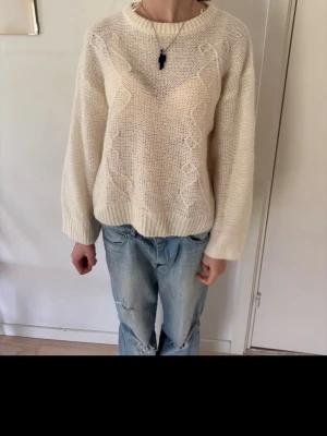 Krämvit stickad tröja med mönster - Mysig krämvit stickad tröja med diskret kabelstickat mönster framtill. Tröjan har rund halsringning, långa ärmar och ribbade muddar vid ärmslut och nederkant. Perfekt för lager på lager och enkel att matcha till jeans.