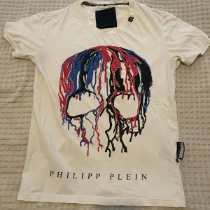 Vit Philipp Plein t-shirt med dödskalle - Vit t-shirt från Philipp Plein med ett stort tryck av en färgglad dödskalle i blått, rött och svart på framsidan. Klassisk passform med korta ärmar och rund hals. Märkesnamnet är tryckt nedtill på framsidan.