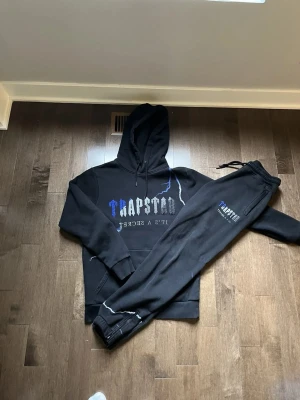 Trapstar svart hoodie och byxa set - Den passar båda M och L i storlek‼️‼️ .Säljer ett svart set från Trapstar med hoodie och matchande byxor. Hoodien har tryck med blå och grå text samt blixtdetaljer och stor ficka framtill. Byxorna har resår i midjan och samma tryck på benet. Mjukt material, troligen bomullsblandning. Perfekt för en streetwear-look.
