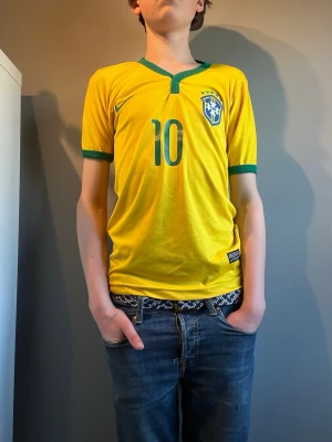 Brasiliansk fotbollströja Nike gul - Säljer en gul Brasilien fotbollströja från Nike med gröna detaljer och nummer 10 på framsidan. Tröjan har korta ärmar, V-ringning och Brasilien-emblem med stjärnor på bröstet. Perfekt för dig som älskar fotboll och vill representera Brasilien.