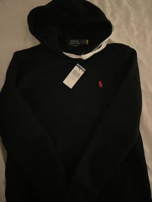 Svart hoodie från Polo Ralph Lauren - Svart hoodie från Polo Ralph Lauren med vit dragsko i huvan och klassisk röd logga broderad på bröstet. Mjuk bomullskänsla och stilren design som passar perfekt till jeans eller joggers.