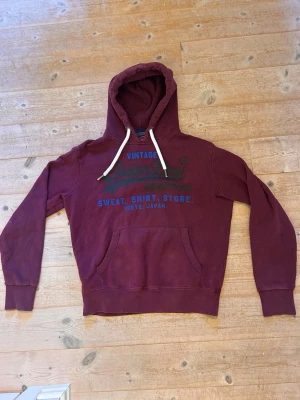 Superdry hoodie - Snygg vinröd hoodie från Superdry med tryck i blått och svart framtill. Klassisk känguruficka och vita dragsnören i huvan. Perfekt för en avslappnad streetwear-look. Mjuk bomullsblandning och bekväm passform.