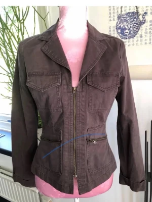 figursydd jacka med dragkedja - Snygg brun/grön   jacka med figursydd passform och markerad midja. Jackan har två bröstfickor med lock, två snedställda fickor med dragkedja och klassisk krage. Stängs med dragkedja framtill och har långa ärmar. Perfekt för en trendig och avslappnad look.