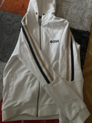 Vit kofta från Hugo Boss - Vit hoodie med dragkedja från Hugo Boss. Tröjan har svarta och beige ränder längs ärmarna, huva med snörning och fickor på sidorna. Boss-logga broderad på bröstet. Perfekt för en sportig och avslappnad stil. Pris kan diskuteras!