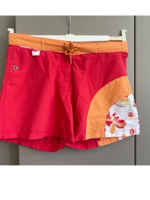 Röda och orangea badbyxor Trespass - Färgglada badbyxor från Trespass i rött och orange med snörning i midjan. Snygg detalj med blommigt mönster på ena sidan och kontrastfärgade paneler. Perfekta för stranden eller poolen. 