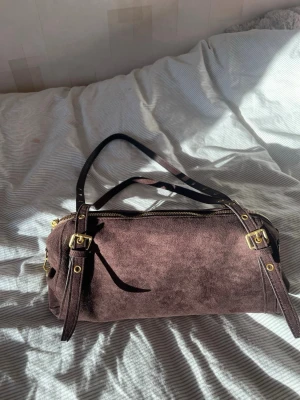 Brun mocka handväska med guldspännen - Säljer en snygg handväska i brun mocka med justerbara svarta axelband och detaljer i guldmetall. Väskan har cylinderformad design och dragkedja upptill. Perfekt för dig som gillar stilrena accessoarer med en lyxig touch.