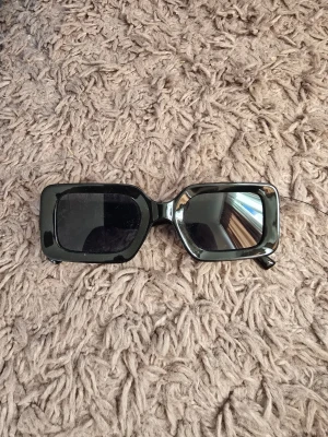 Svarta rektangulära solglasögon - Snygga solglasögon med rektangulära svarta bågar och mörka glas. Metallbågarna ger en clean och modern look som passar till de flesta outfits. Perfekta för soliga dagar när du vill ha en stilren accessoar.