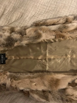 Beige pälsväst från Hollies - Snygg och fluffig pälsväst från Hollies i beige och bruna toner. Västen har ett lyxigt, mjukt foder och är tillverkad i äkta päls. Perfekt för att lyfta din outfit med en trendig touch.