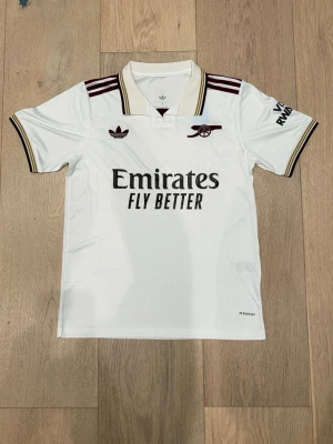 Vit Arsenal Adidas matchtröja - Snygg vit Arsenal matchtröja från Adidas med bruna detaljer och klubbens logga på bröstet. Tröjan har korta ärmar, krage med ränder och tryckta sponsordetaljer. Perfekt för fotbollsträning eller att bära på läktaren.