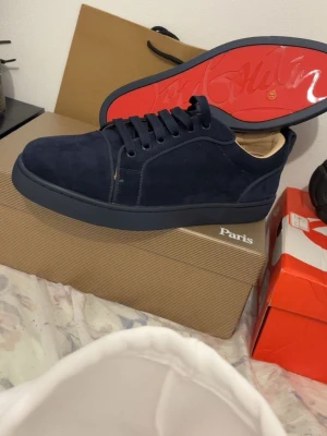 Mörkblå sneakers från Christian Louboutin - Snygga mörkblå sneakers i mocka från Christian Louboutin med klassisk röd sula och lågt skaft. Skorna har matchande mörkblå snörning och en stilren design som sticker ut med den ikoniska röda undersidan.