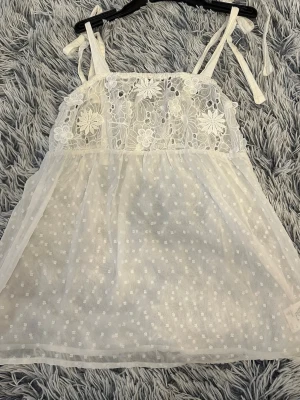 Vit spetslinnetopp med broderi - Söt vit linnetopp med tunna axelband och knyt. Överdelen har blommigt spetsbroderi och nederdelen är i tunn mesh med små prickar. Perfekt för sommarens varma dagar. Pris går att ändra