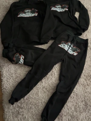 Svart loungewear-set Trapstar Shooters - Svart loungewear-set från Trapstar med trycket 'London Shooters' och pantermotiv på både tröja och byxor. Setet består av sweatshirt och mjukisbyxor med resår i midjan och muddar vid benslut. Perfekt för chill dagar hemma.