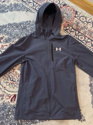 Mörkblå vindjacka från Under Armour - Snygg mörkgrå vindjacka från Under Armour med huva och dragkedja framtill. Jackan har en bröstficka med dragkedja och logga på vänster sida. Perfekt för blåsiga dagar och sportiga outfits.