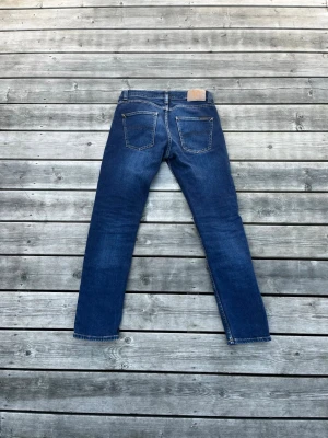 Nudie Jeans - Mörkblå jeans från Nudie Jeans med rak passform och klassisk femficksdesign. Jeansen har knappgylf och kontrastsömmar, tillverkade i denim av bomull. Perfekt för en stilren och avslappnad look.